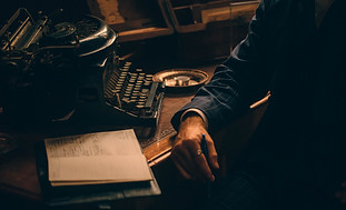 A dimly lit scene with an old-fashioned typewriter on a wooden desk. A person in a suit is partially visible, holding a pen and resting one hand on a notebook. The ambiance suggests vintage or historical themes.