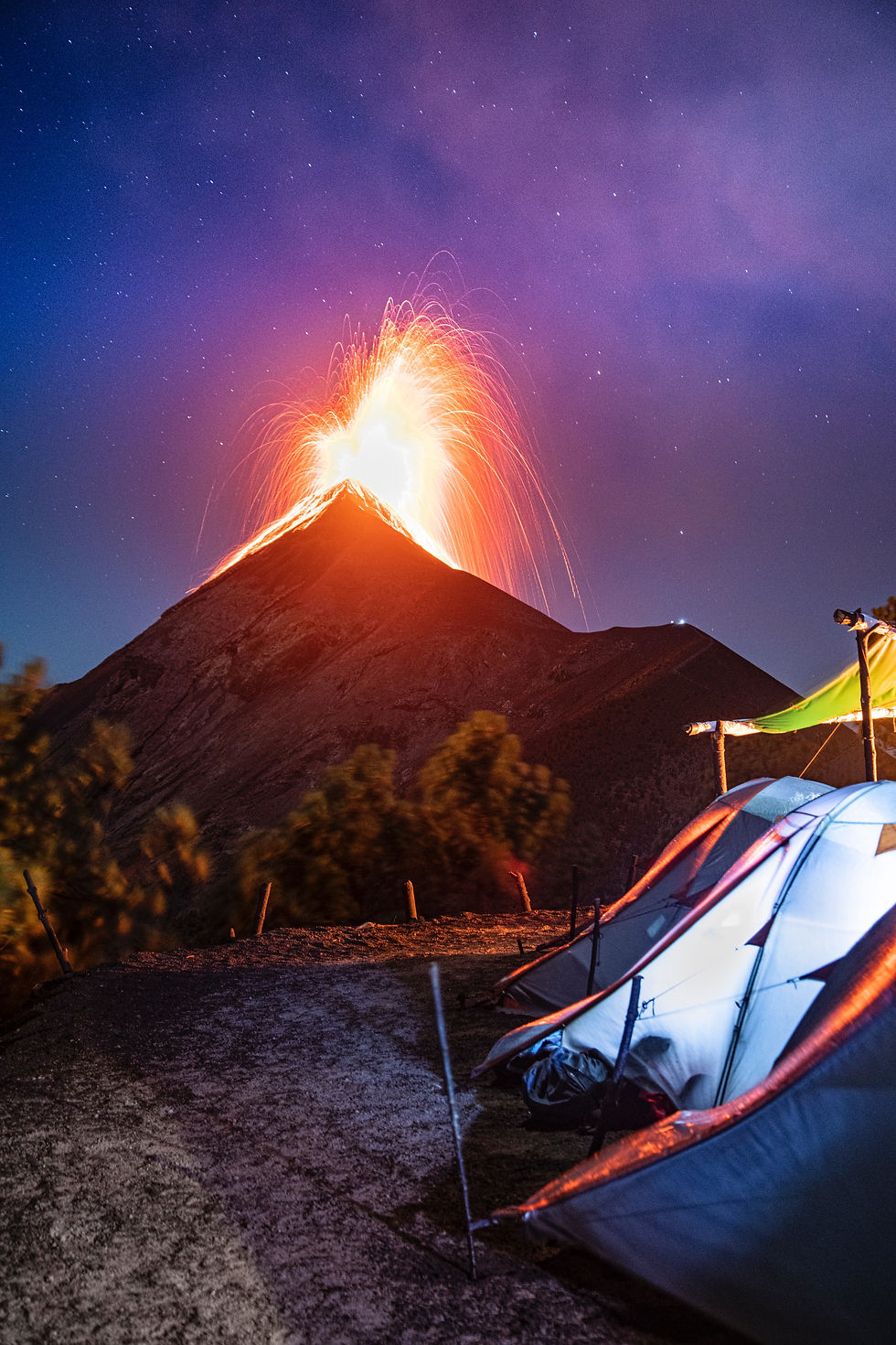 Volcano erupting at night, casting fiery sparks against a starry sky. Tents are set up in the foreground, creating a dramatic scene.