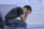 Boy in black shirt and jeans sits on a chair, head in hands, appearing distressed. White door and tiled floor in the background.