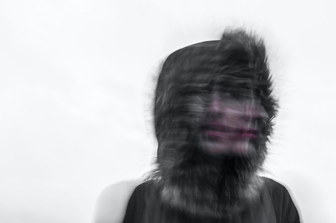 Blurred person in a hood with fur trim against a cloudy sky. The face appears distorted, creating an eerie, abstract effect.