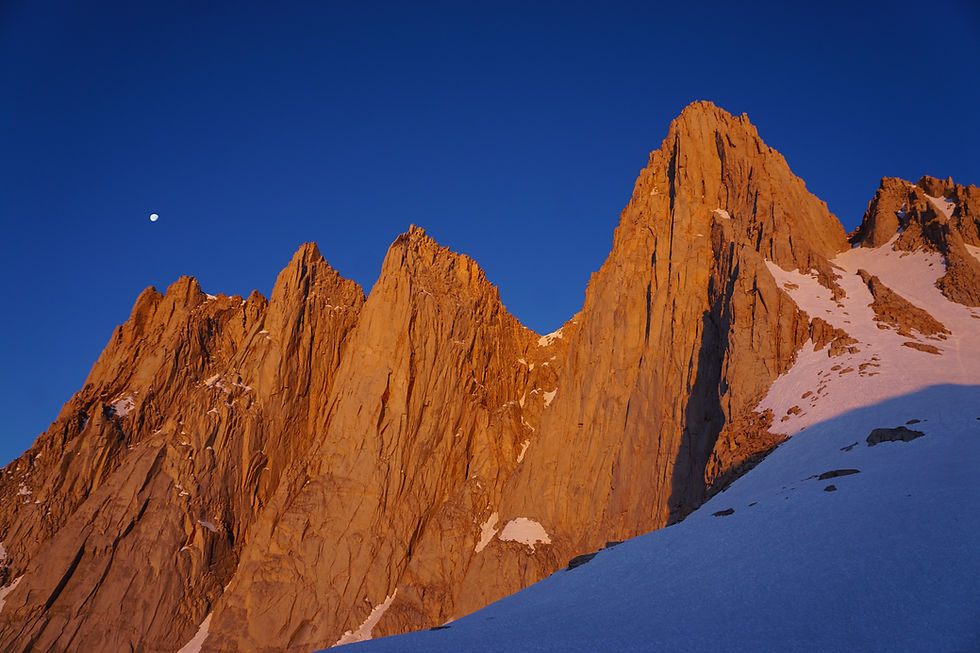 Jagged Mount Whitney peaks lit by sunset, with a deep blue sky and a white moon, casting shadows on snow-covered slopes.