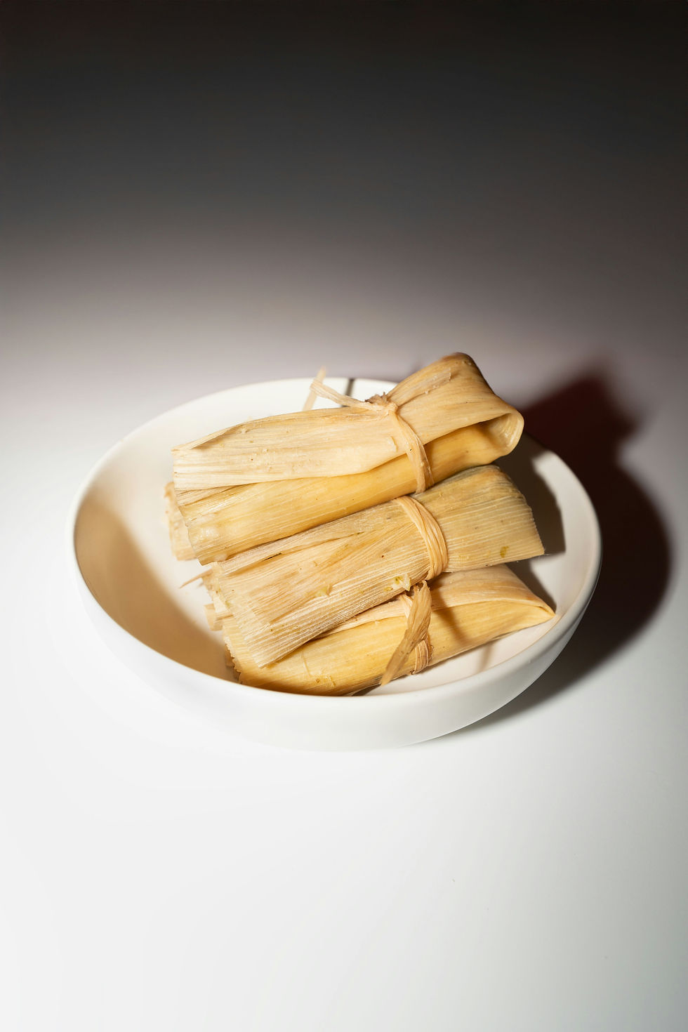 Three tamales wrapped in corn husks sit in a white bowl on a white surface under bright light.