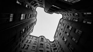 Black-and-white view from a courtyard looking up at tall apartment buildings framing a bright sky, with curved facades and many windows.