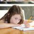 Young girl with brown hair leans over a notebook, drawing with a yellow pencil at a wooden table in a bright home, focused.