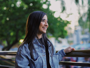 Smiling woman in a denim jacket leans on a railing outdoors, with a soft green, sunlit background.