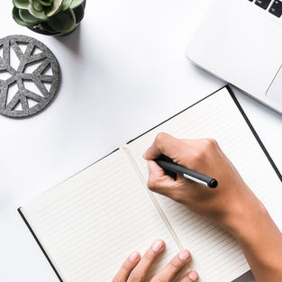 An open notebook with a plant and laptop in each corner, a persons hand is holding a pen above the notebook about to write.