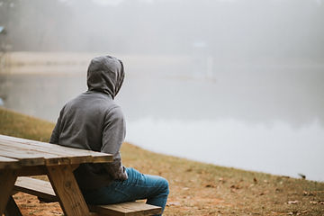 Hooded person sits on a wooden picnic bench beside a foggy lake, facing the water in a quiet, reflective mood.