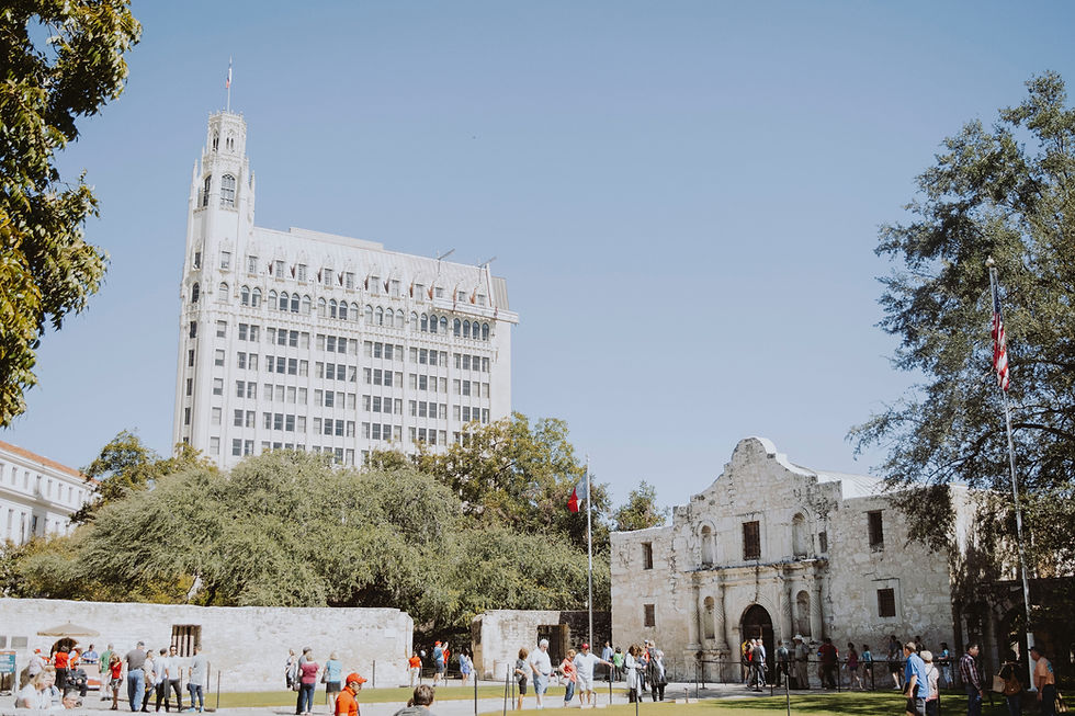 A sunny day at the Alamo in San Antonio, Texas, with visitors gathered outside the historic mission, and a tall building in the background.