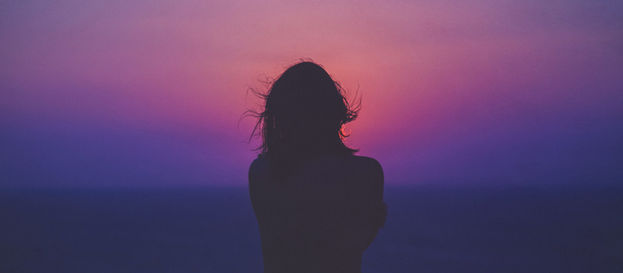 A lone silhouette stands against a mesmerizing sunset, her hair gently ruffled by the breeze, immersed in the tranquil hues of purple and pink.