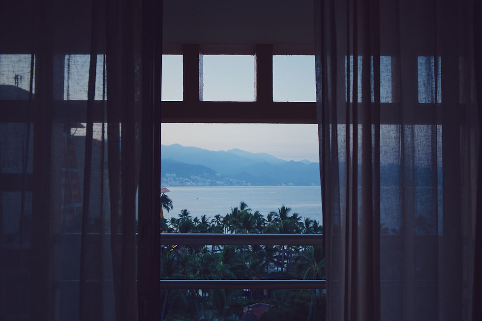 An opened window with dark curtains on either side looks out onto a tropical landscape with the ocean, rolling hills, and palm trees.