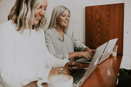 zwei Frauen mit blonden Haaren und weißen Blusen sitzen an ihren Laptops und lachen