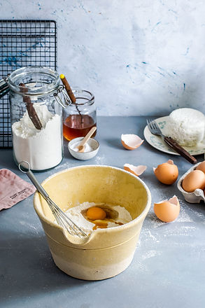 Bowl with flour, egg, and whisk on a gray countertop. Nearby: cracked eggshells, a jar of flour, honey, and cheese on a plate. Baking setup.