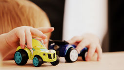 Child playing with toy cars on a wooden table. One car is bright yellow and another is blue. The background is softly blurred.