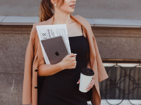 Woman in a tan coat and black dress holds a tablet, newspaper, and coffee cup. Background: gray stone wall and metal railing.