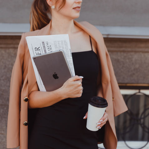 Woman in a tan coat and black dress holds a tablet, newspaper, and coffee cup. Background: gray stone wall and metal railing.