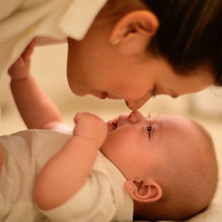 Woman gently touches noses with smiling baby, both in white outfits on a soft, beige background, creating a warm, tender moment.