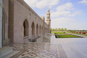 Sultan Qaboos Grand Mosque Muscat Oman. Elegant mosque exterior with arched windows and tall minaret. Forecourt features patterned tiles. Palms and flowerbeds add color under blue skies.