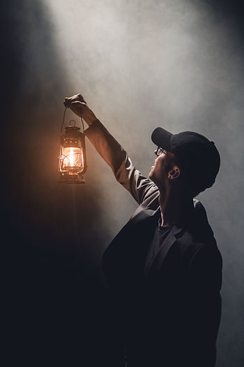 Hombre con gorra y chaqueta sosteniendo una lámpara de aceite encendida en la oscuridad, representando la búsqueda de luz y dirección espiritual en medio de la oscuridad.