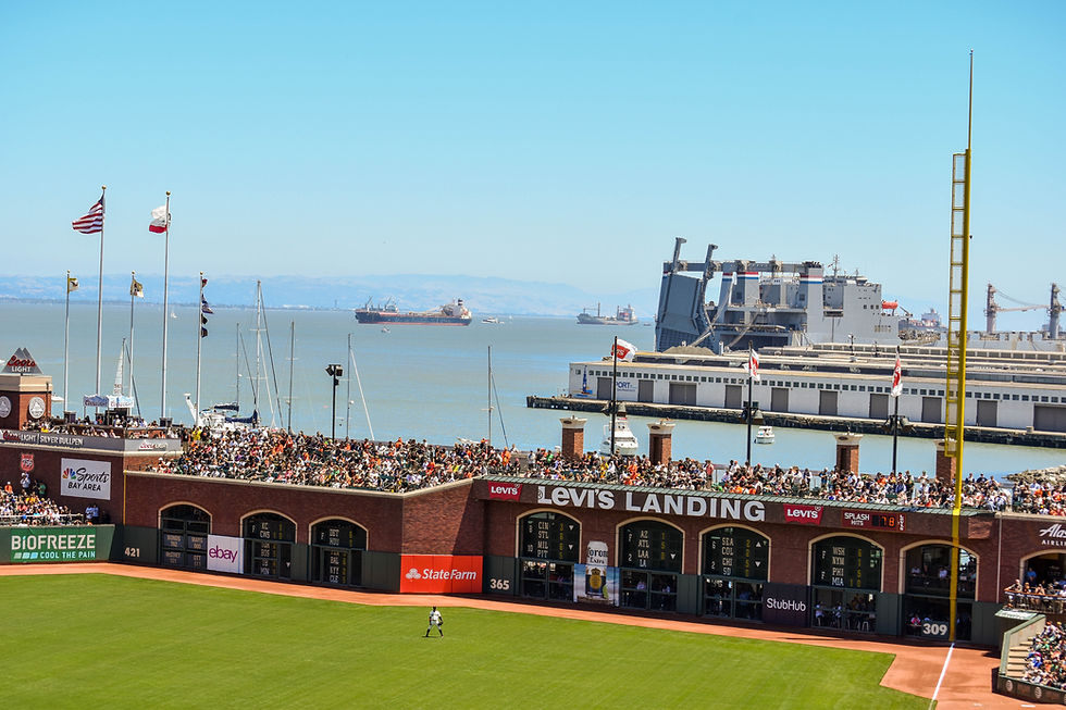 Crowd at Levi's Landing watches a baseball game by the waterfront. Flags fly, ships sail in the distance, clear blue sky.