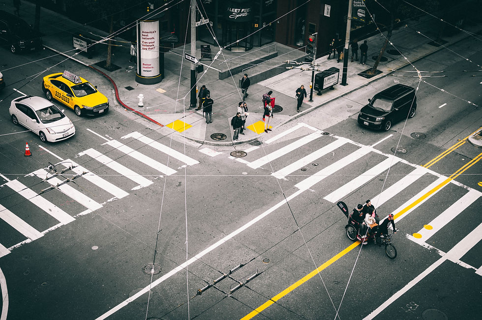 The image shows an aerial view of a busy urban intersection with multiple crosswalks. Pedestrians are crossing the street in various directions, and a few people are gathered near the center, one of whom is on a