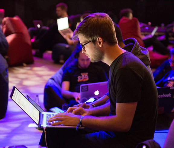 A young man wearing eye glasses and a black t-shirt works with his Mac - more people in the background