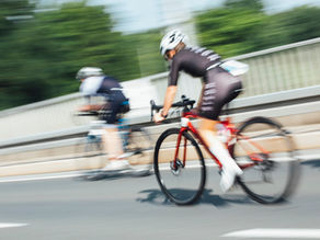 Blurred cyclists race across a bridge, one on a red bike in black kit and white helmet, with green trees behind.