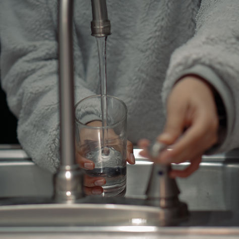 A person in a gray fleece fills a glass with water at a kitchen sink, adjusting the tap under dim lighting.