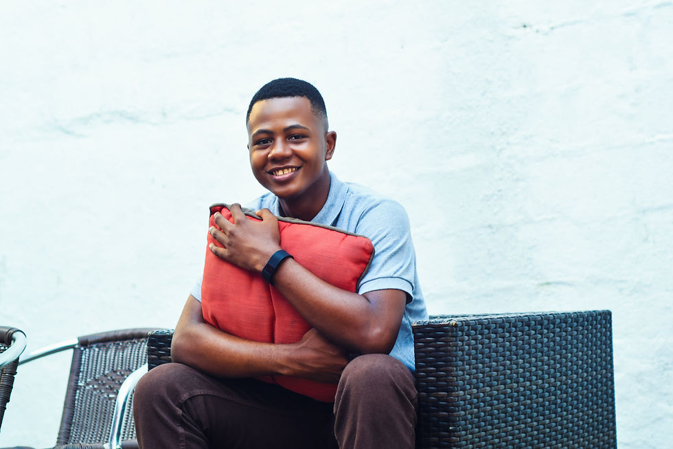 Man smiling, seated on wicker chair, hugging a red pillow against a light blue wall. Casual attire, relaxed mood, and simple setting.