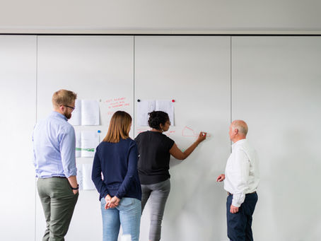 Four people collaborate at a whiteboard, one drawing a red diagram. Papers are pinned, and notes are visible. Office setting.