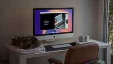 Cat sleeping on folded blanket beside an iMac showing We Design Websites, with keyboard and headphones on a tidy desk by a window