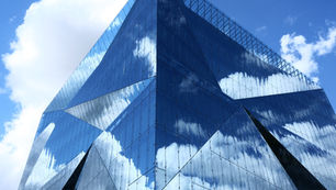 A modern glass building with angular architecture reflecting blue sky and white clouds, creating a seamless and futuristic look.