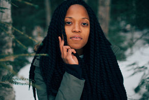Woman with long braids touches her face, standing in a snowy forest. She wears a green jacket, with a calm expression.