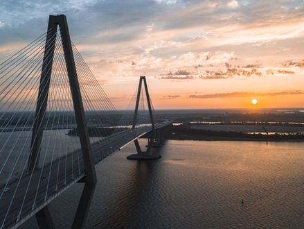 Suspension bridge over a calm river at sunset, with vibrant orange and pink skies. A serene and peaceful atmosphere prevails.