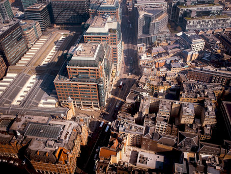 Aerial view of a cityscape with modern and older buildings, roads, and parked vehicles. Shadows suggest bright sunlight. Urban, bustling.