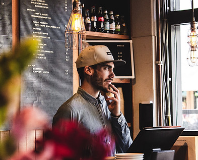A cashier in a brown hat standing at a cash register in a cafe. Menu items are displayed behind him.