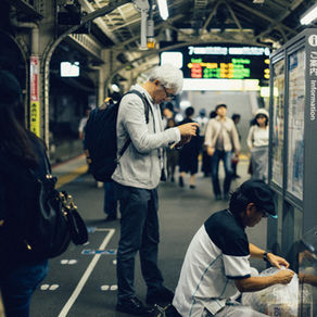 駅のホームで、白髪の男性がスマホを操作し、清掃員がゴミを整理。背景に人々が歩き、案内板や電光掲示板が見える。