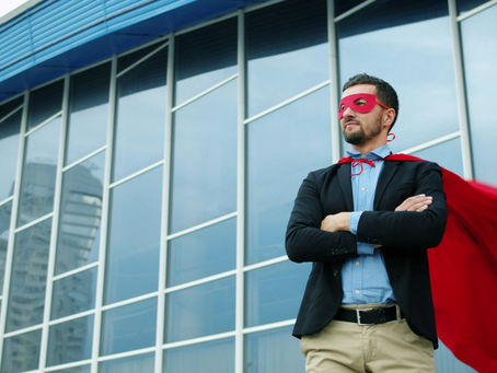 Devant un immeuble vitré, un homme en veste de costume foncée, chemise bleue pantalon beige, les bras croisés porte un masque et une cape rouge et regarde au loin