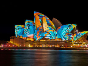 Sydney Opera House illuminated with vibrant blue and orange butterfly patterns at night, reflected in the harbor, creating a festive mood.