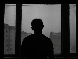 Silhouette of person gazing out a window, urban buildings visible through fog. Black and white, creating a somber, reflective mood.