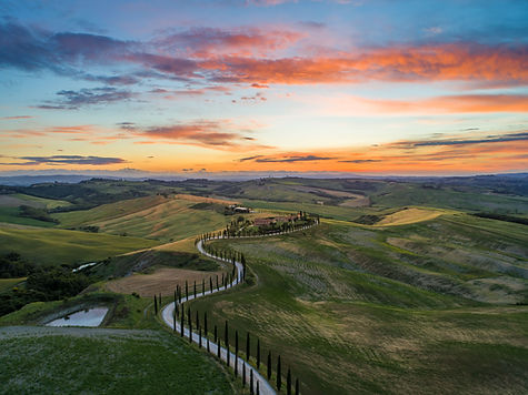 Far-away view of a Tuscan villa at sunset overlooking rolling hills and a tree-lined road in the countryside.