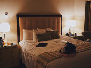 Cozy bedroom with a beige tufted headboard bed, lamps on nightstands, and items like a laptop and water bottle. Soft lighting creates a calm mood.
