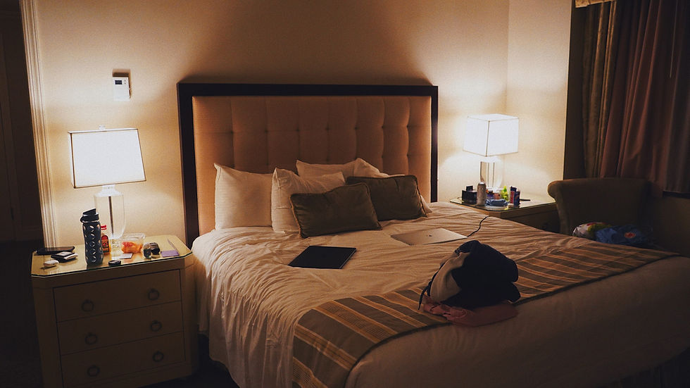 Cozy bedroom with a beige tufted headboard bed, lamps on nightstands, and items like a laptop and water bottle. Soft lighting creates a calm mood.