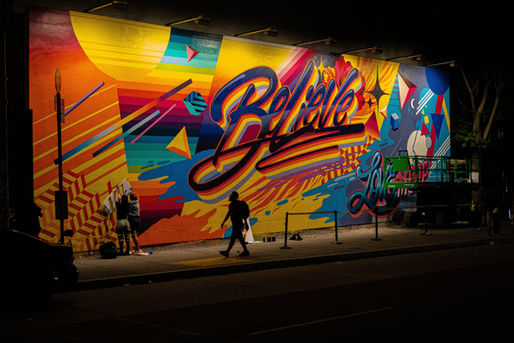 Colorful mural with "Believe" text, vibrant geometric patterns. Two people working on it. Nighttime setting, silhouette of passerby.