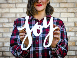 Woman in a red plaid shirt holds a white "joy" sign against a brick wall. She has a subtle smile and wears bold red lipstick.