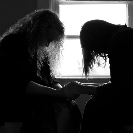 Two silhouetted women sit facing each other, heads bowed and hands clasped, in front of a bright window in a quiet black-and-white room.
