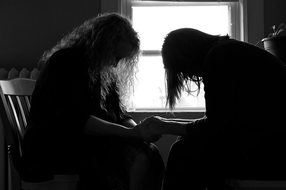 Two silhouetted women sit facing each other, heads bowed and hands clasped, in front of a bright window in a quiet black-and-white room.