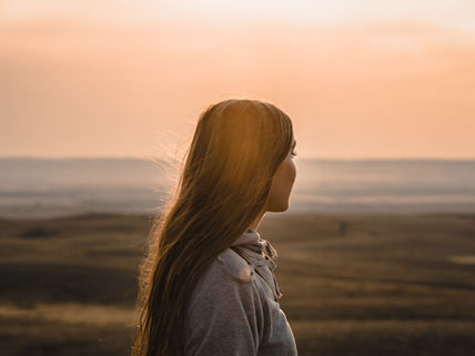 Young woman in profile at a windy seaside sunset, long hair glowing in warm light, gazing over the calm horizon