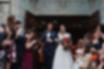 Bride and groom exit a stone building, smiling among cheering guests who blow bubbles. Bride holds a vibrant bouquet. Joyful atmosphere.