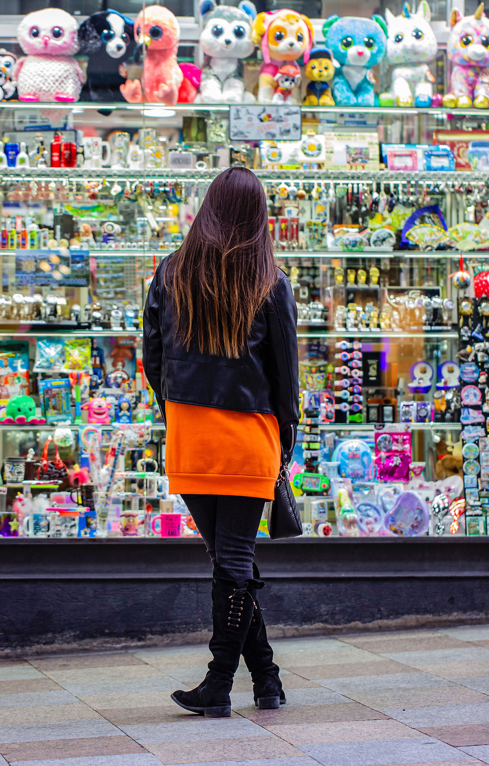 Woman in orange hoodie and black boots stands facing a vibrant store display with colorful stuffed animals and trinkets.
