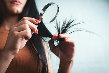 A woman holding and inspecting the dry, split ends of her hair against a neutral background.
